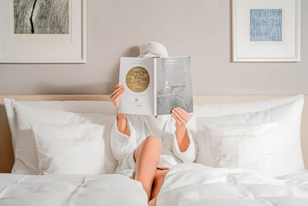 Woman in a robe on a hotel bed holding a magazine up, for an article about relaxing travel